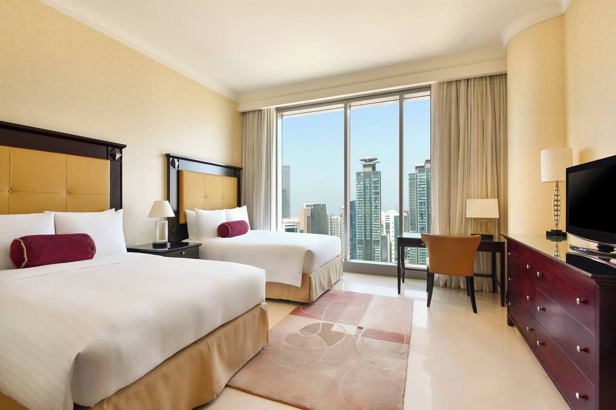 A hotel room with two beds, a TV, a desk, and a view of the city.
