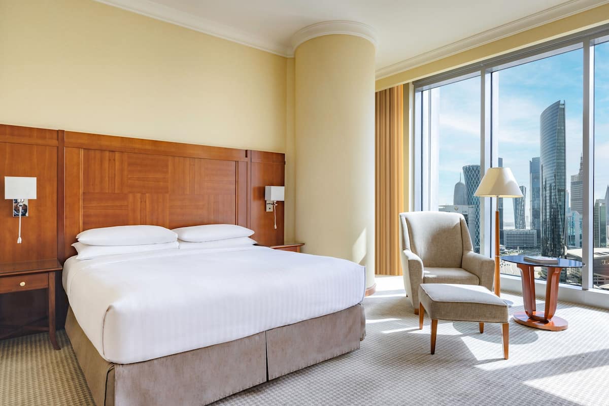 A hotel room with a large bed, a chair, a small table, and a view of the cityscape through large windows.