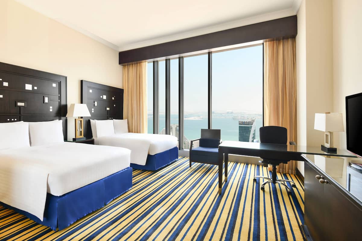 A hotel room with two beds, a desk, a chair, a TV, and a large window with a city view.