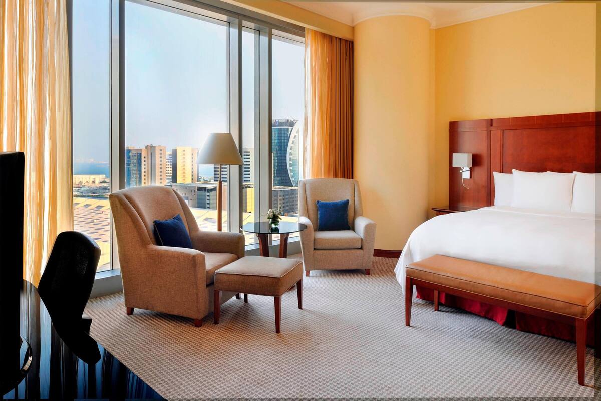 A hotel room with a large bed, two armchairs, a small table, and a view of the cityscape.