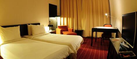 A hotel room with two beds, a desk, a chair, a TV, and a large window with curtains.