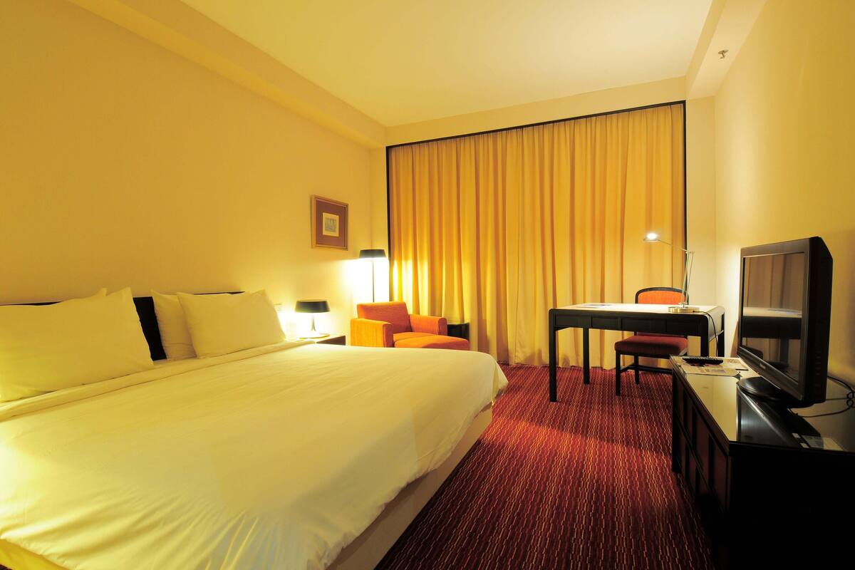 Habitación de hotel con una cama grande, un escritorio con silla, televisión y ventana con cortinas.