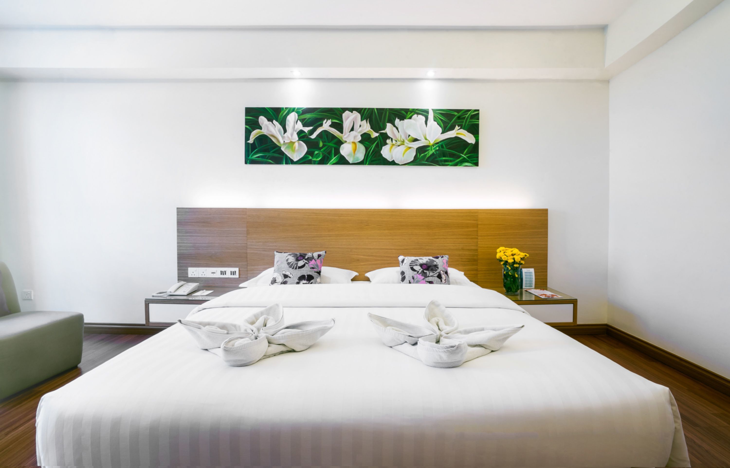 Habitación de hotel con una cama grande, cabecera de madera, mesita de noche con teléfono y un cuadro de flores colgado en la pared.