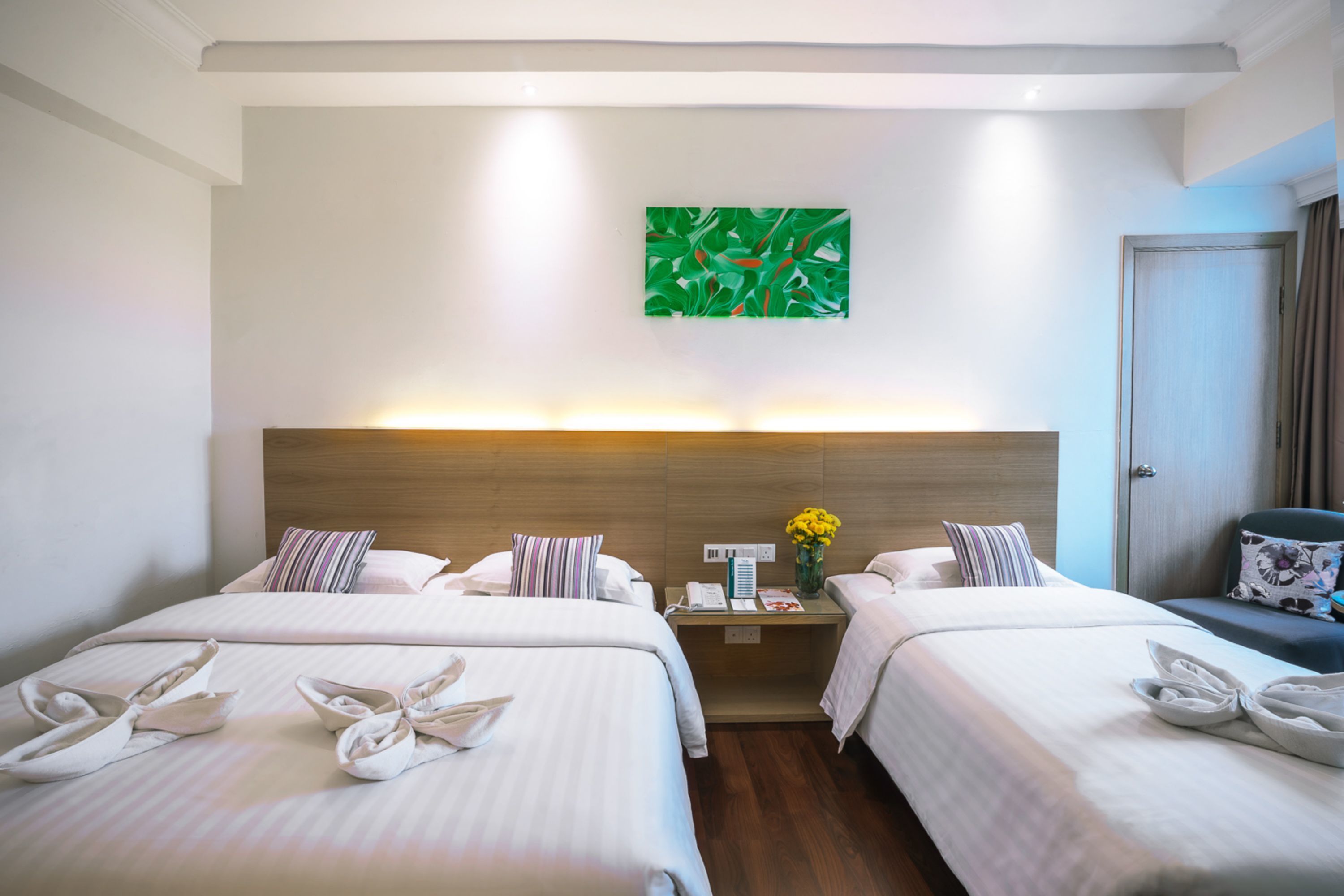 A hotel room with two beds, a nightstand with a lamp, a green abstract painting on the wall, and a door to the right.