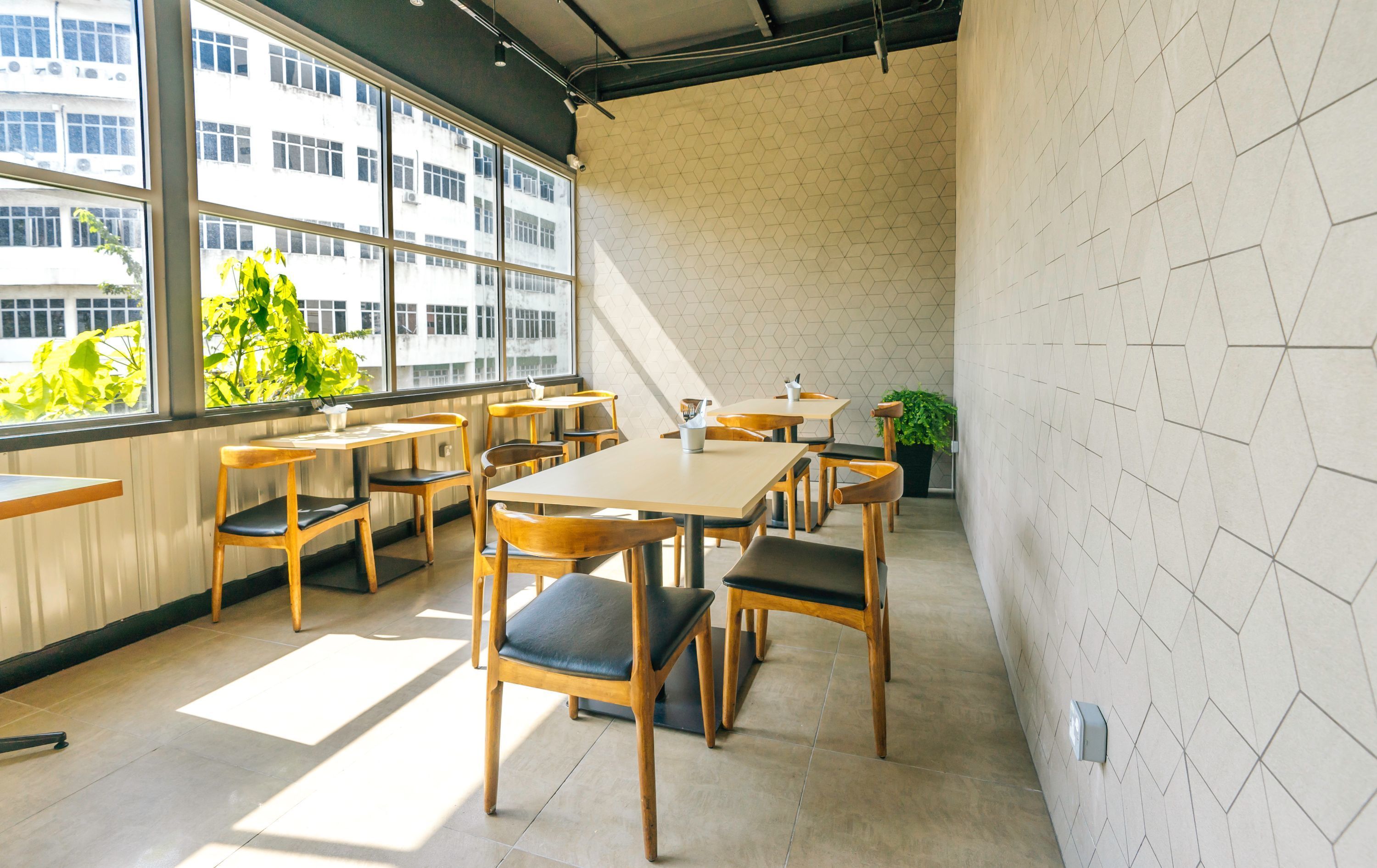 Un comedor moderno con mesas y sillas de madera, ventanas amplias y un diseño geométrico en la pared.