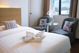 A hotel room with a neatly made bed, two armchairs, a small table with a vase, and a window with a city view.