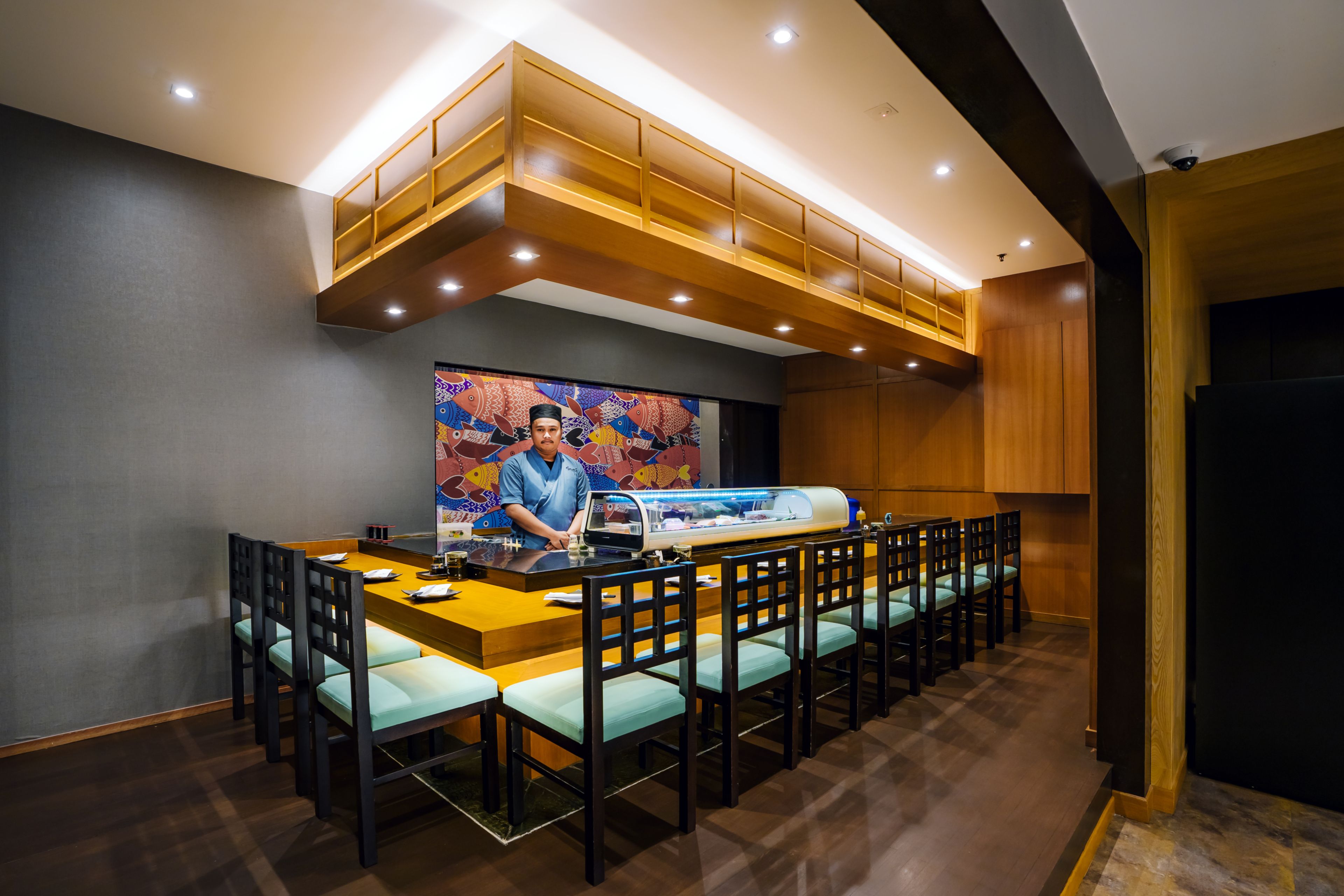 A restaurant with a long counter, multiple chairs, and a chef at the front.