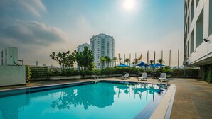 Una piscina con agua azul cristalina, rodeada de sillas de descanso y sombrillas, con un paisaje urbano y edificios altos al fondo.