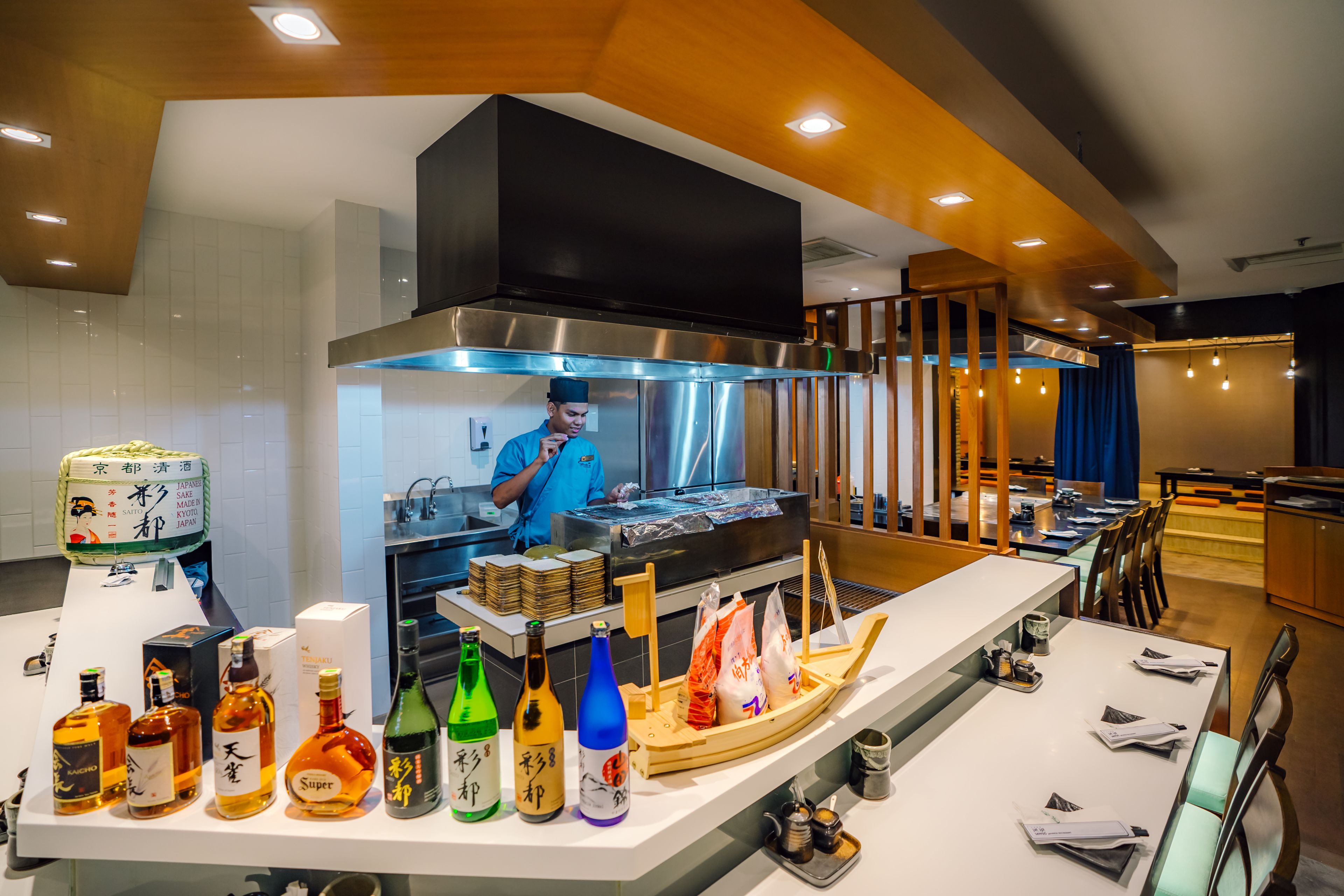 Un intérieur de restaurant moderne, avec un chef en train de préparer des plats, une variété de bouteilles d’alcool exposées et une salle à manger propre et bien aménagée.