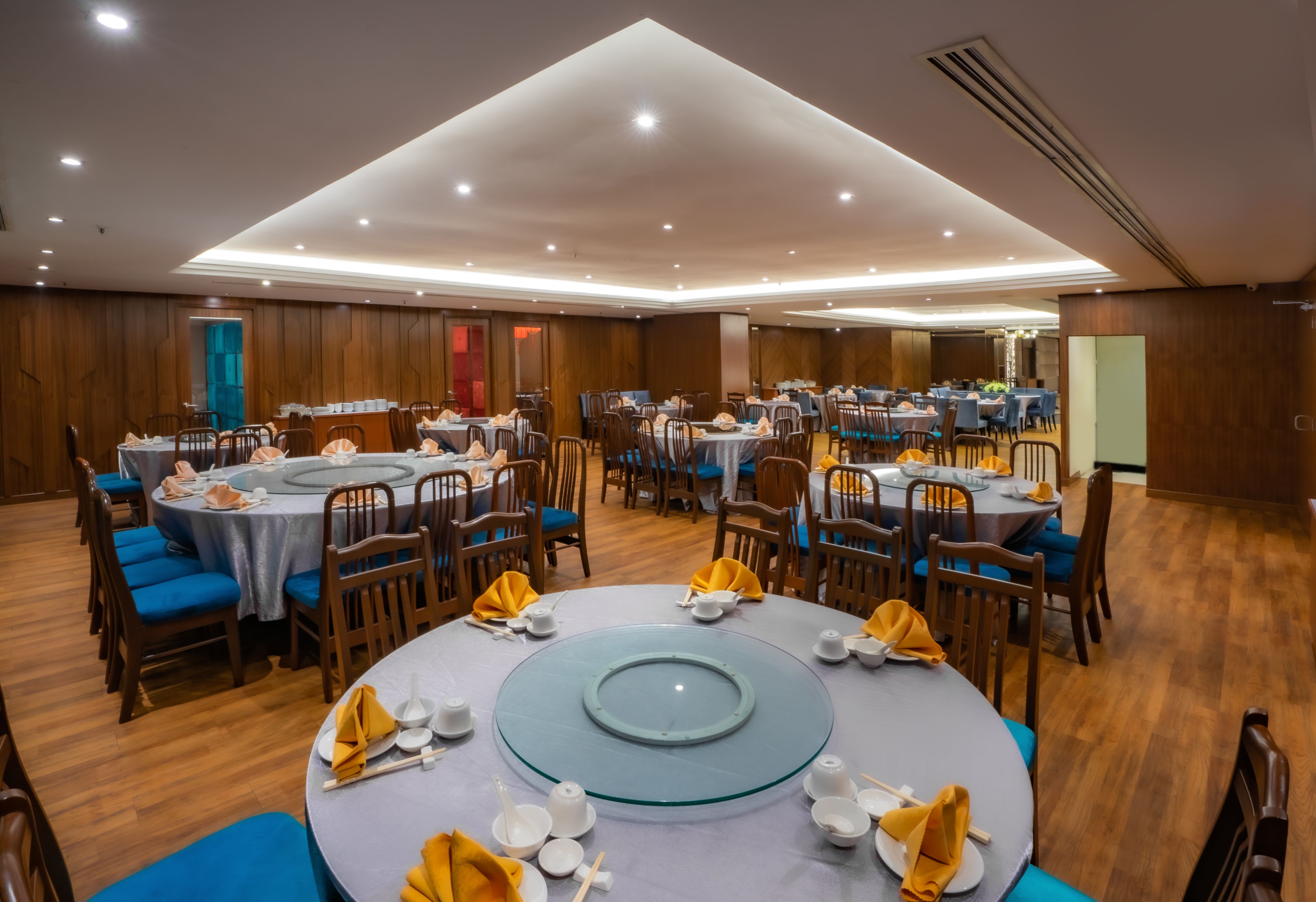 Un comedor espacioso con múltiples mesas redondas, cada una con platos blancos, tazones y servilletas amarillas. La habitación presenta paneles de madera y un techo alto con iluminación empotrada.