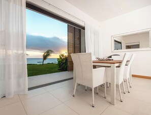 A dining area with a view of the ocean, white chairs, a wooden table, and a window with blinds. - Cap Ouest by Dream Escapes (Flic-en-Flac)