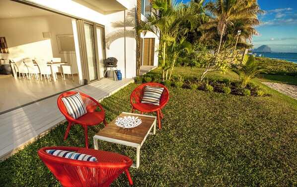 A modern outdoor seating area with red chairs and a wooden table, a covered patio with a dining set, and a view of the ocean and palm trees. - Cap Ouest by Dream Escapes (Flic-en-Flac)