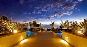 Un sentiero di legno conduce a una piscina con vista sull'oceano e un cielo al tramonto.