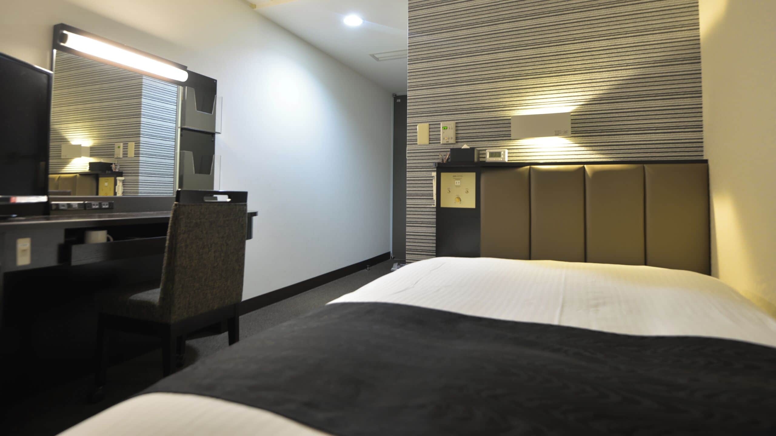 Habitación de hotel con cama, escritorio, silla, espejo y cabecera con iluminación integrada.