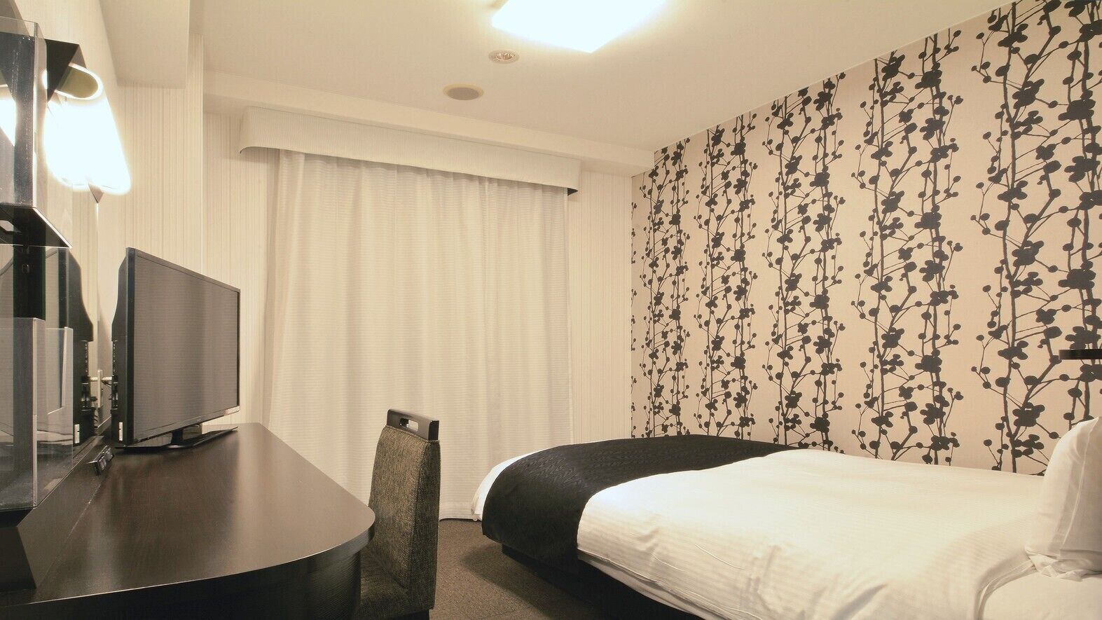 Et hotelværelse med en seng, et skrivebord med en computer, et fjernsyn og et vindue med gardiner.