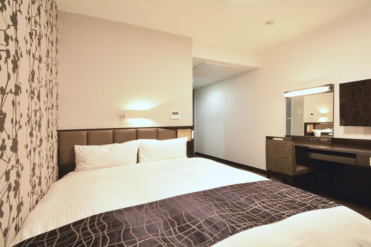 Habitación de hotel con cama, escritorio, silla, espejo y refrigerador pequeño.