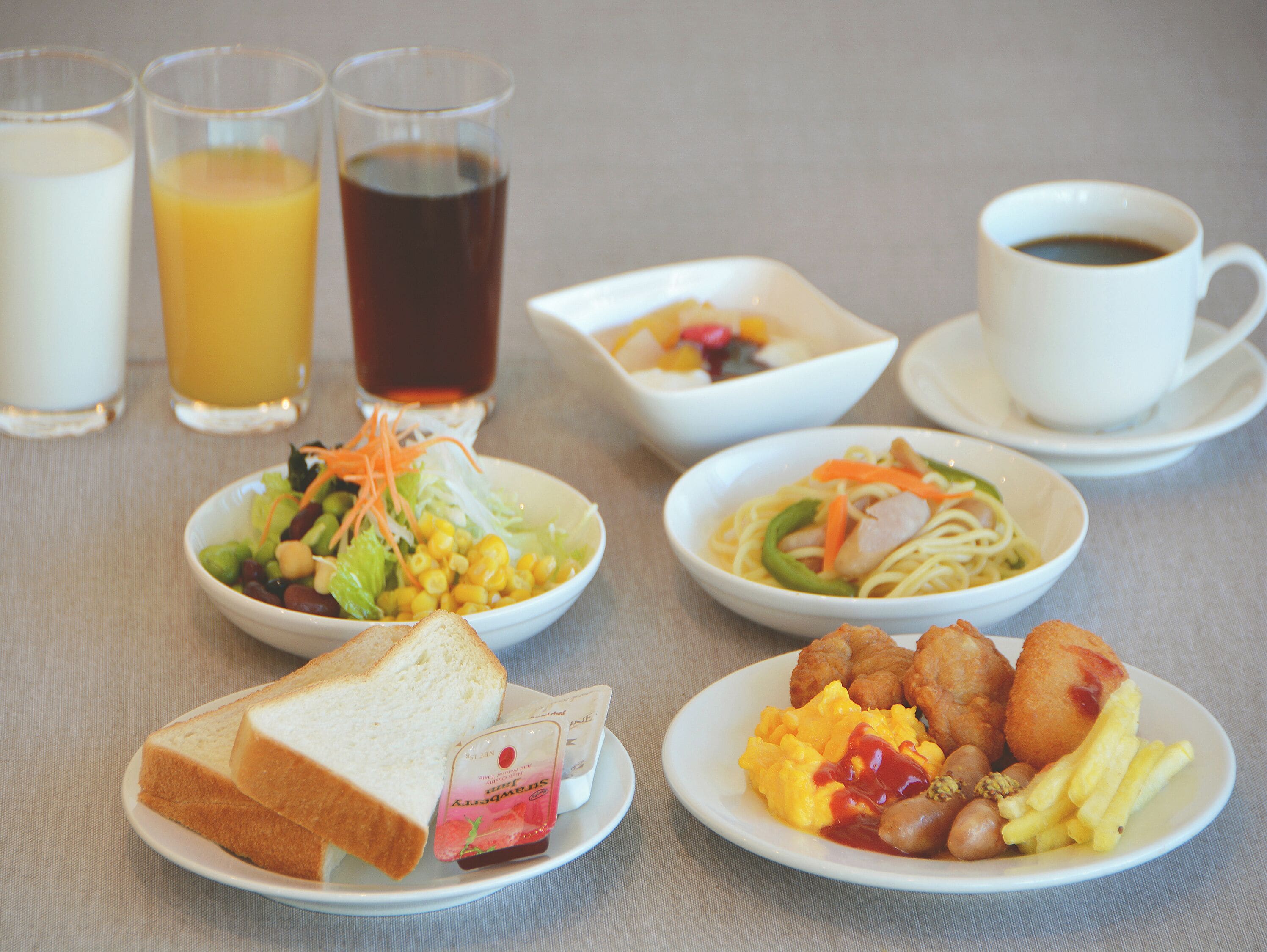 A breakfast setting with various dishes, including toast, scrambled eggs, sausages, and a bowl of salad. There are also two glasses of juice and a cup of coffee.