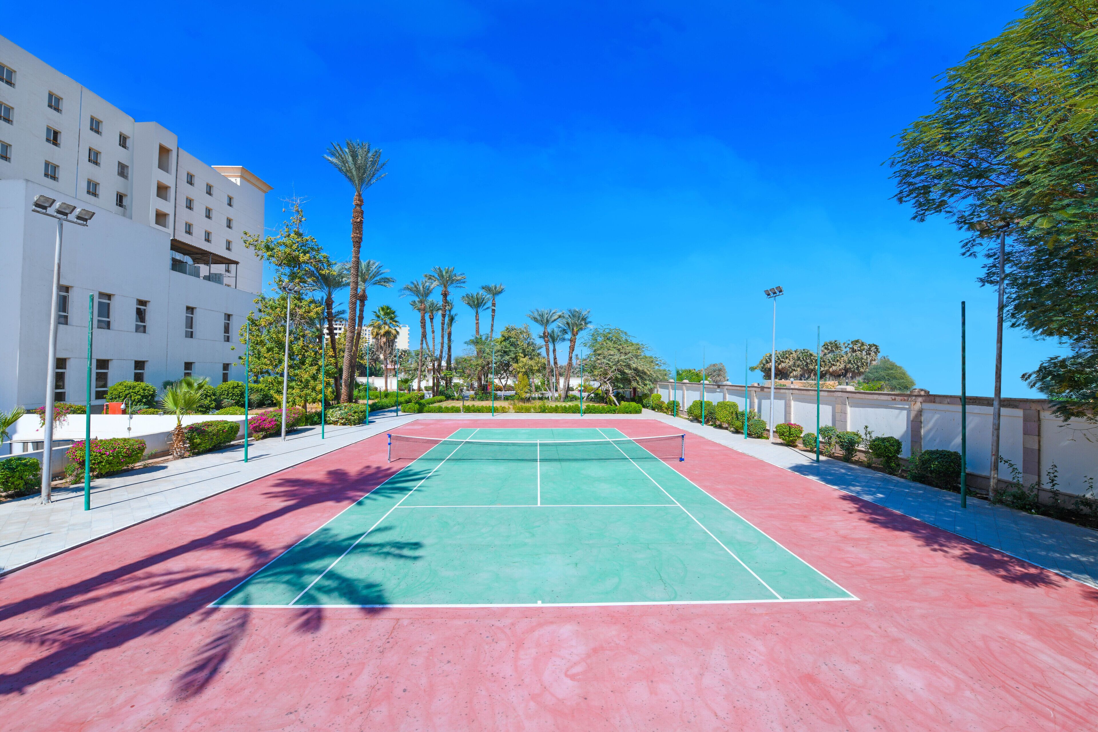 An outdoor tennis court with a red and green surface, surrounded by palm trees and a white building.
