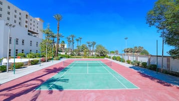 An outdoor tennis court with a red and green surface, surrounded by palm trees and a white building.