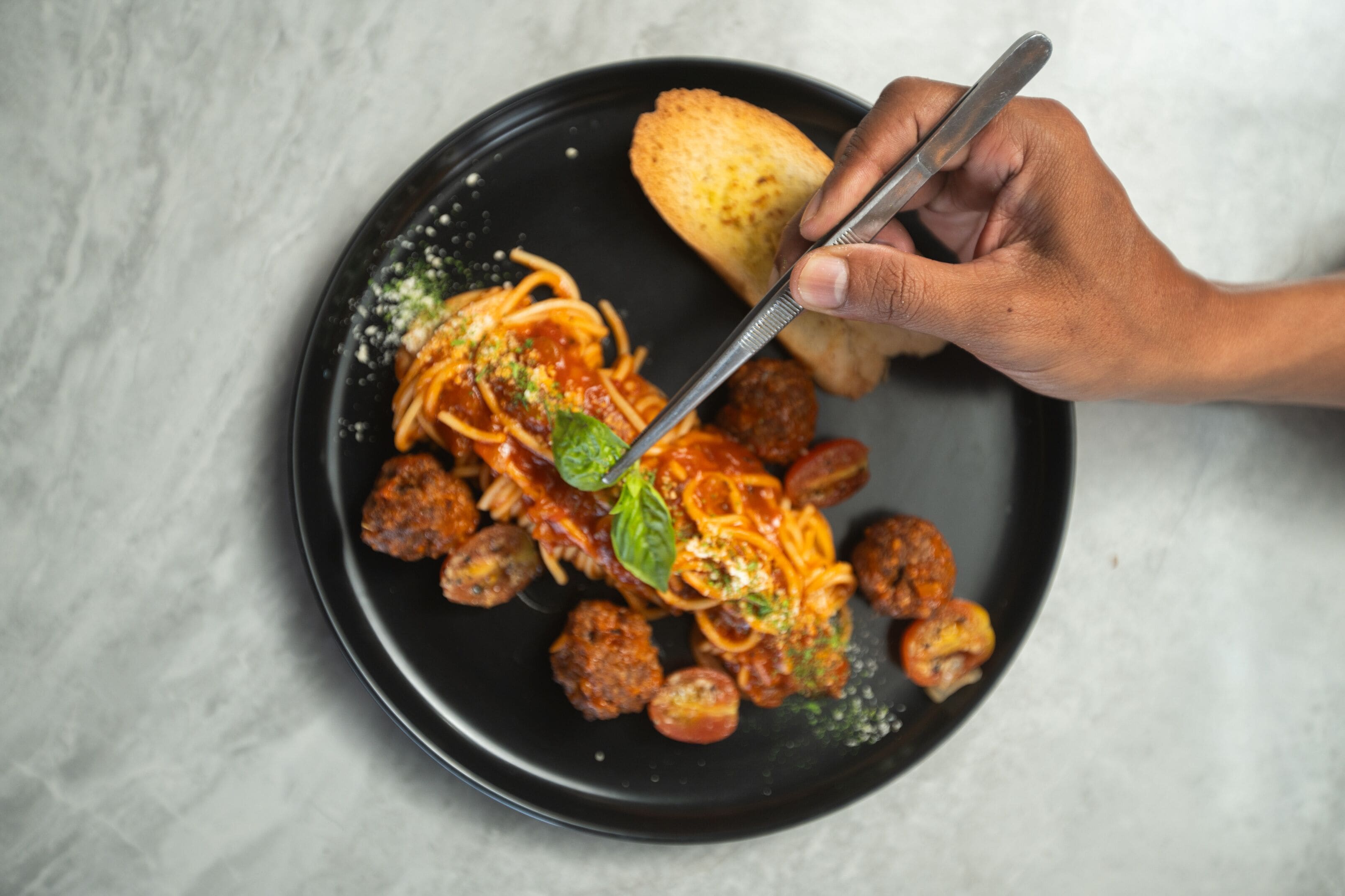 A black plate with spaghetti, meatballs, and a piece of bread on a marble surface.