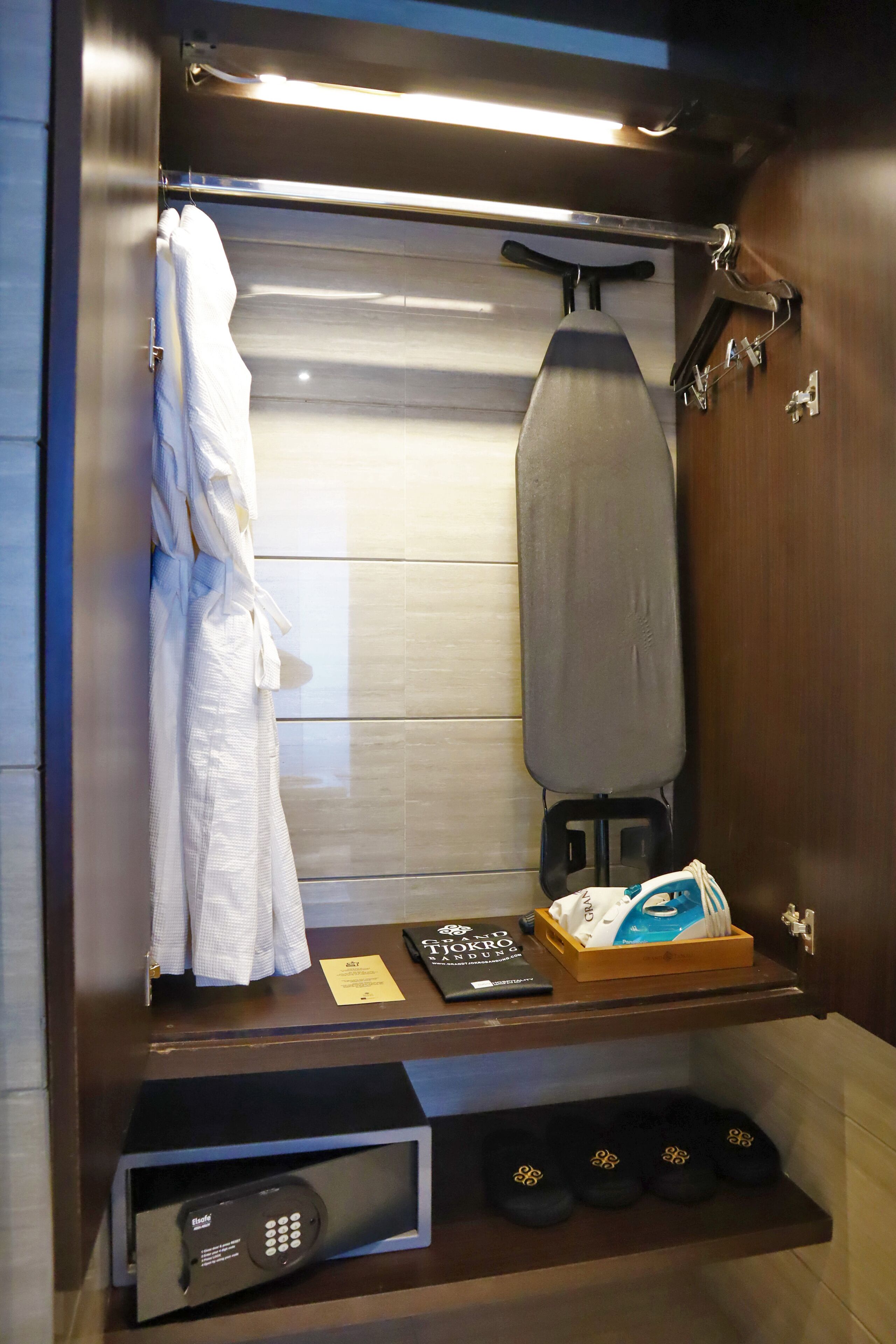 A compact bathroom with a towel rack, an ironing board, a shoe rack, and a small electronic device on a shelf.