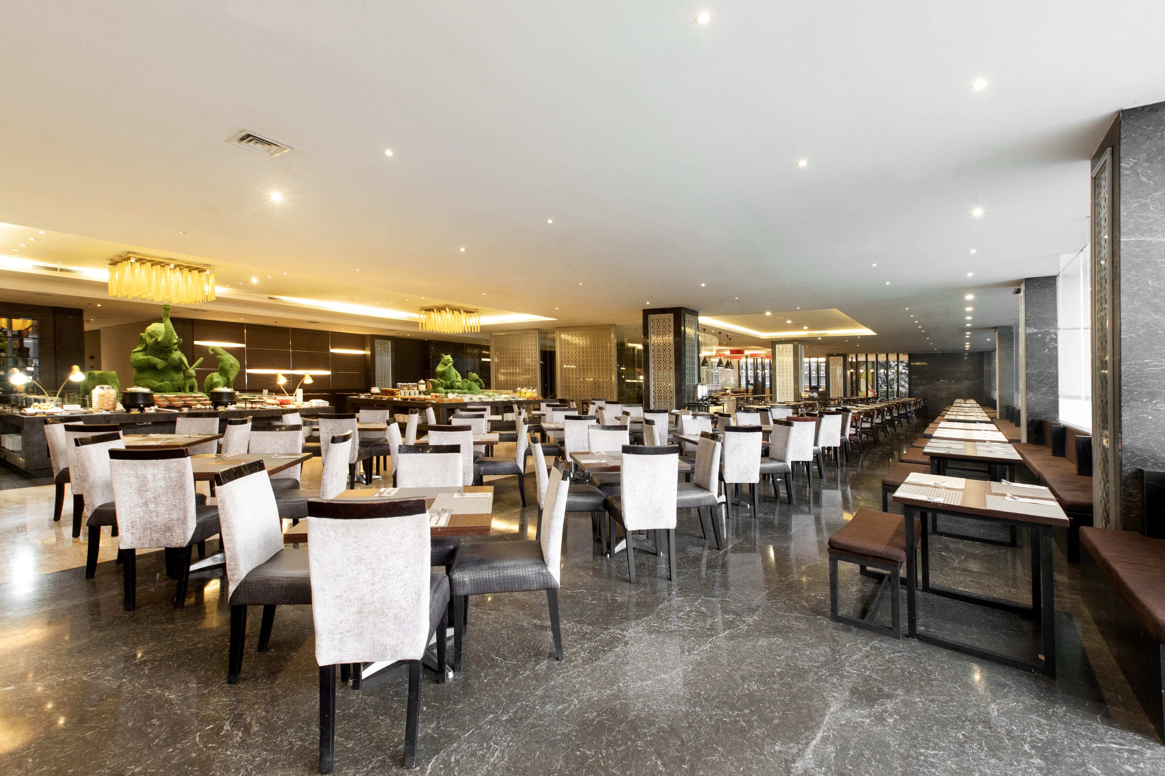 A spacious hotel dining area with multiple tables and chairs, a central buffet, and a modern chandelier.