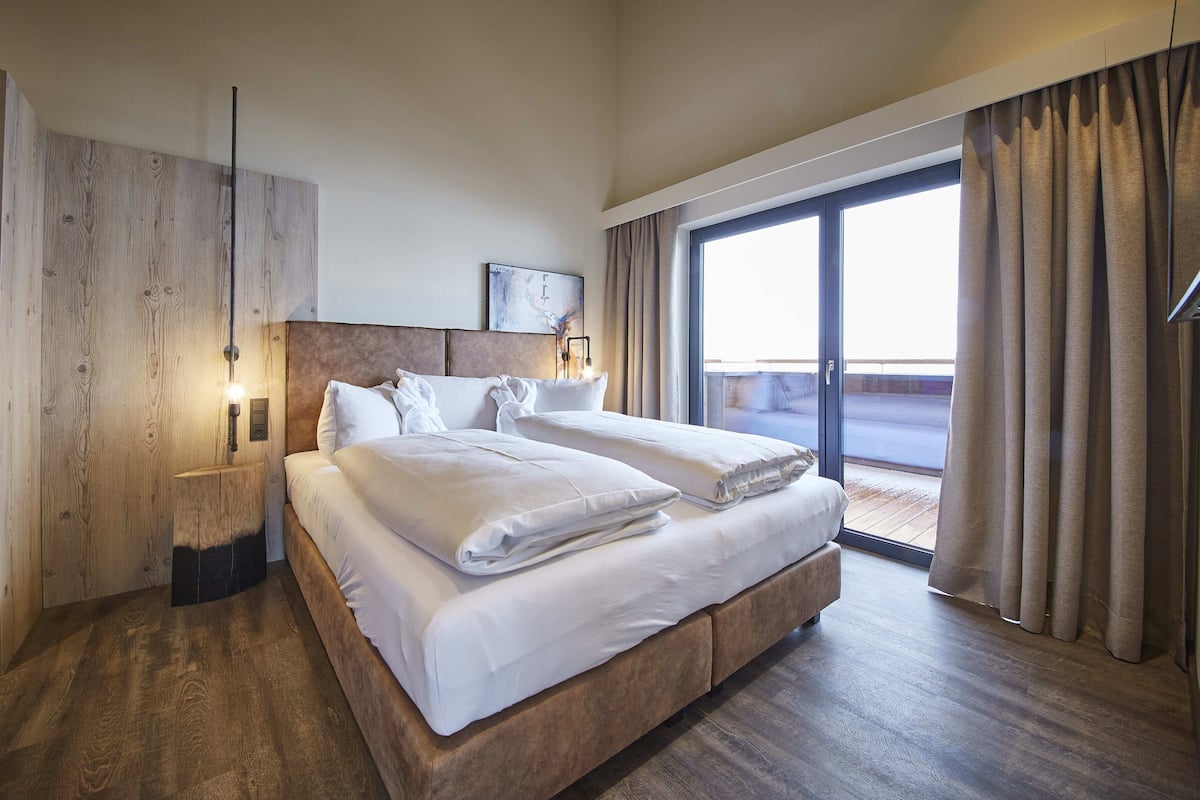 Een hotelkamer met een groot bed, een houten hoofdbord, een nachtkastje, een wandlamp en uitzicht op de omgeving door een glazen schuifdeur.