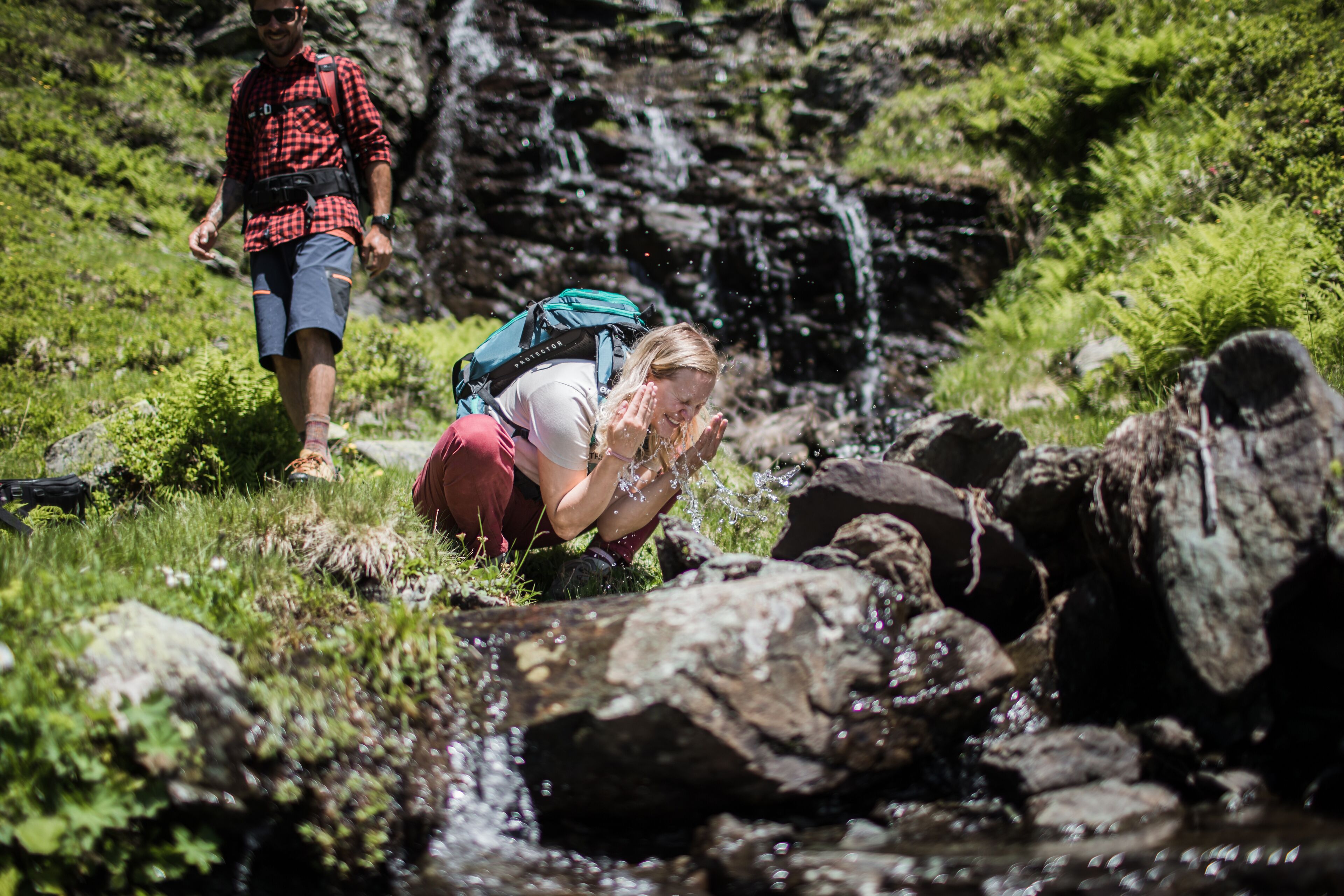 A person crouching by a stream with rocks and greenery around. Another person is walking in the background.