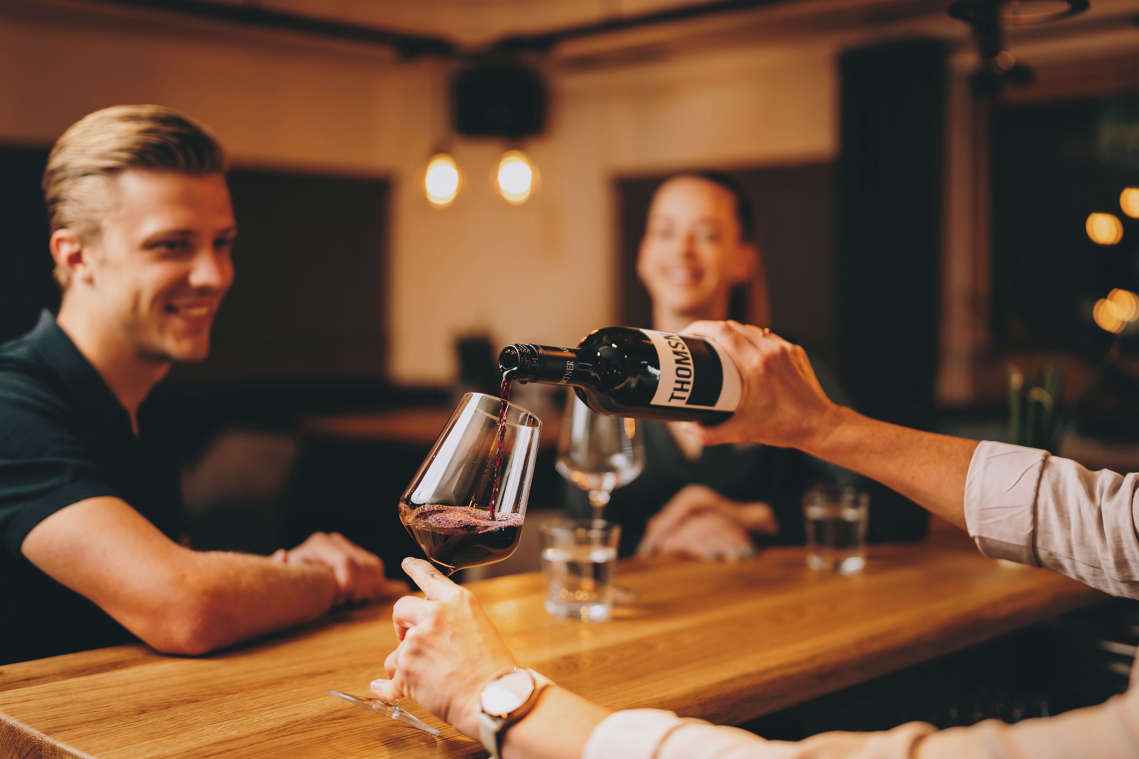 A person pouring wine into a glass at a wooden table in a dimly lit indoor setting.