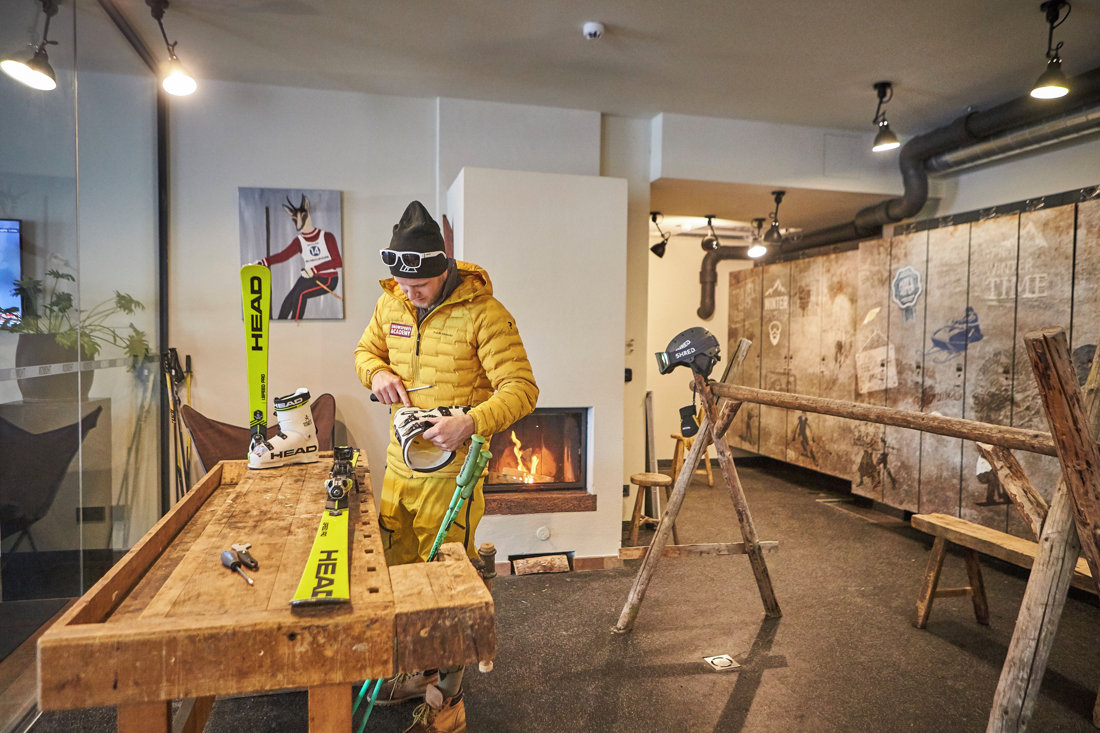 A person in a yellow jacket adjusting ski bindings on a wooden table with ski equipment.