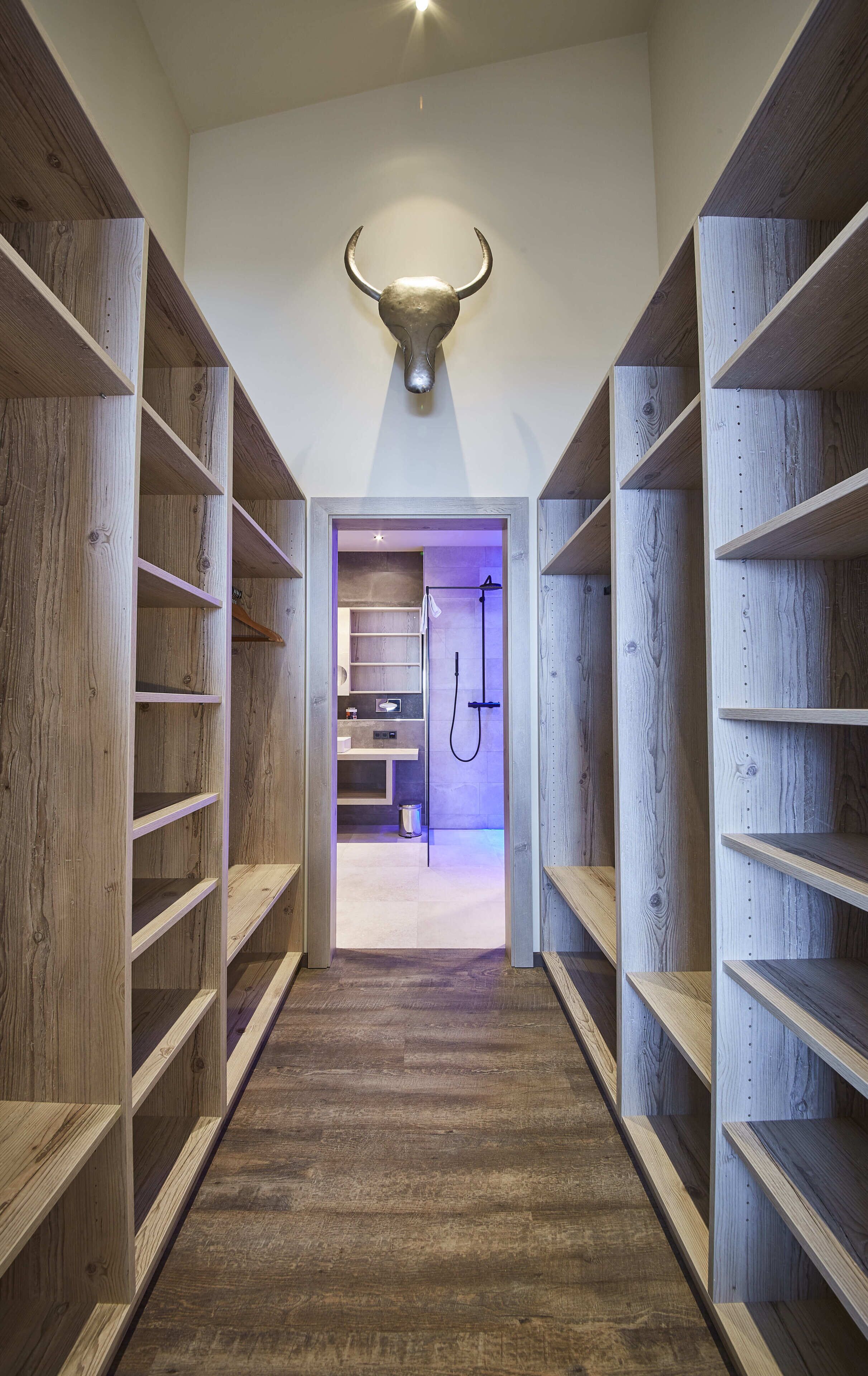 A narrow hallway with wooden shelving on both sides, leading to a bathroom with a shower and a sink.