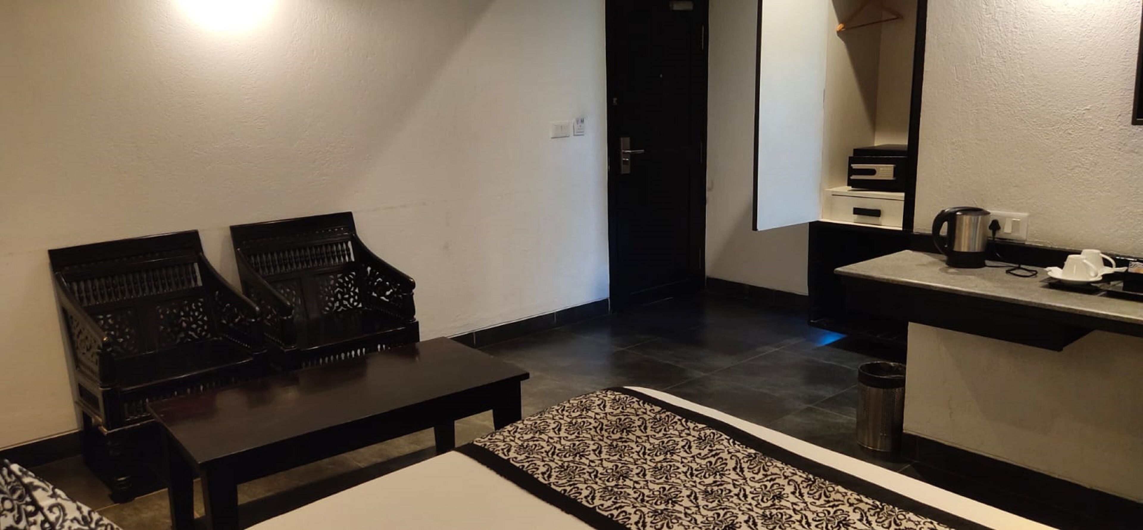 A hotel room with a dark wooden bench, a small table, a patterned rug, a sink with a kettle, and a mirror.