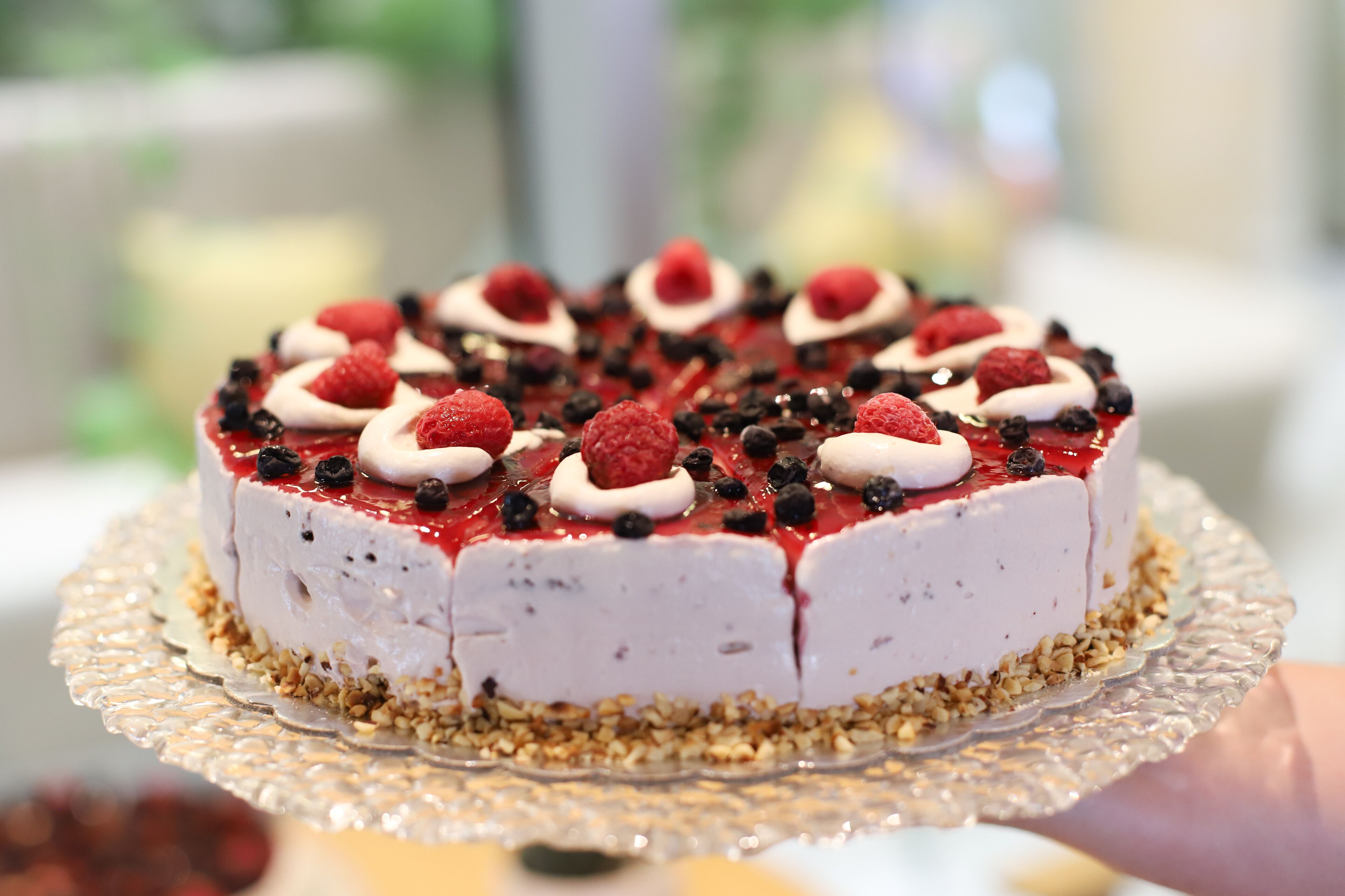 Een ronde taart met witte glazuur, frambozen en chocoladestukjes op een glazen taartplateau.
