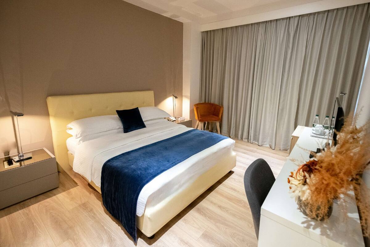A hotel room with a bed, bedside table, chair, and a desk with a vase.