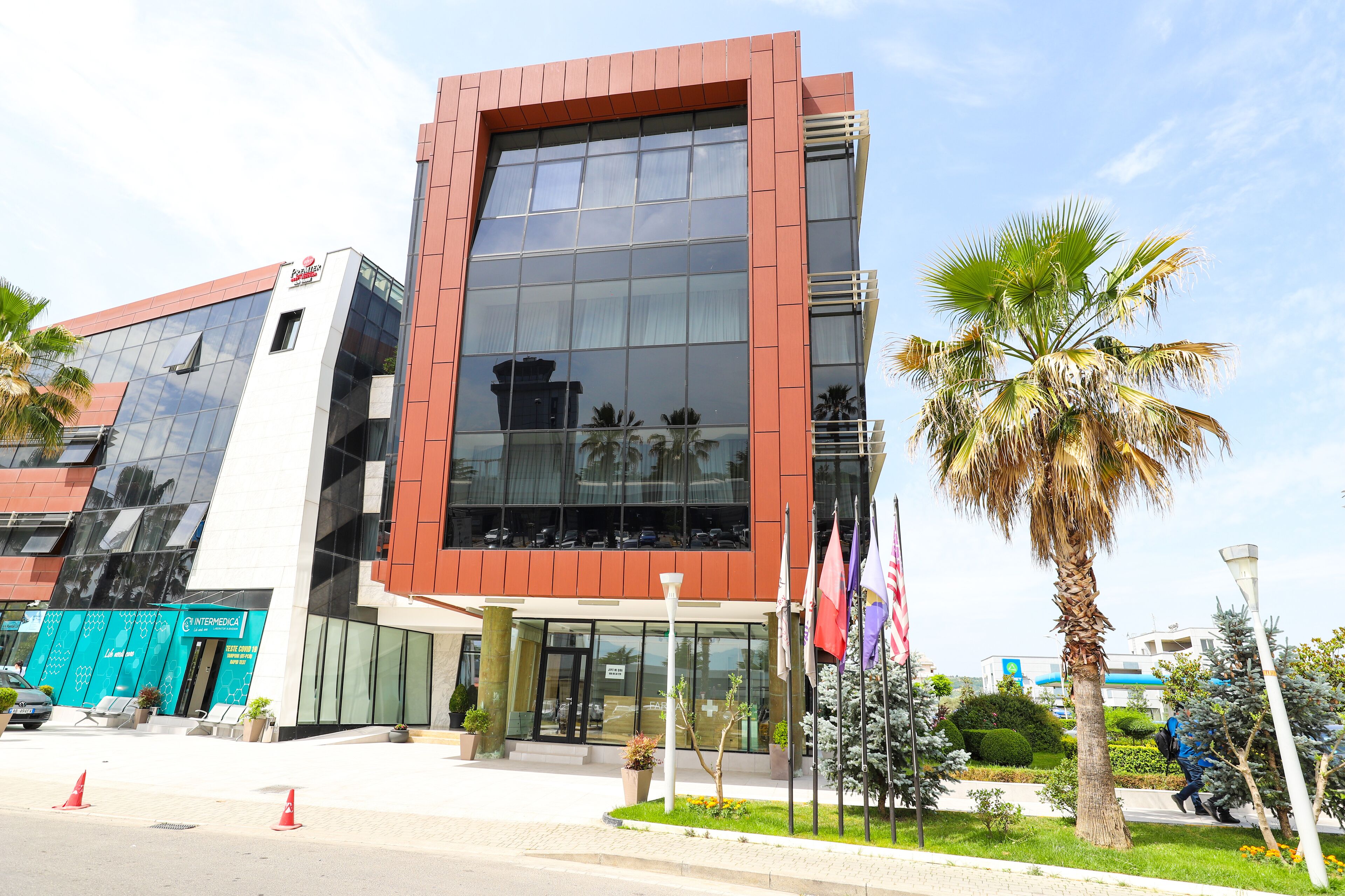 Un edificio moderno con fachada de vidrio, exterior rojo y blanco, y un pequeño jardín con palmeras y banderas.