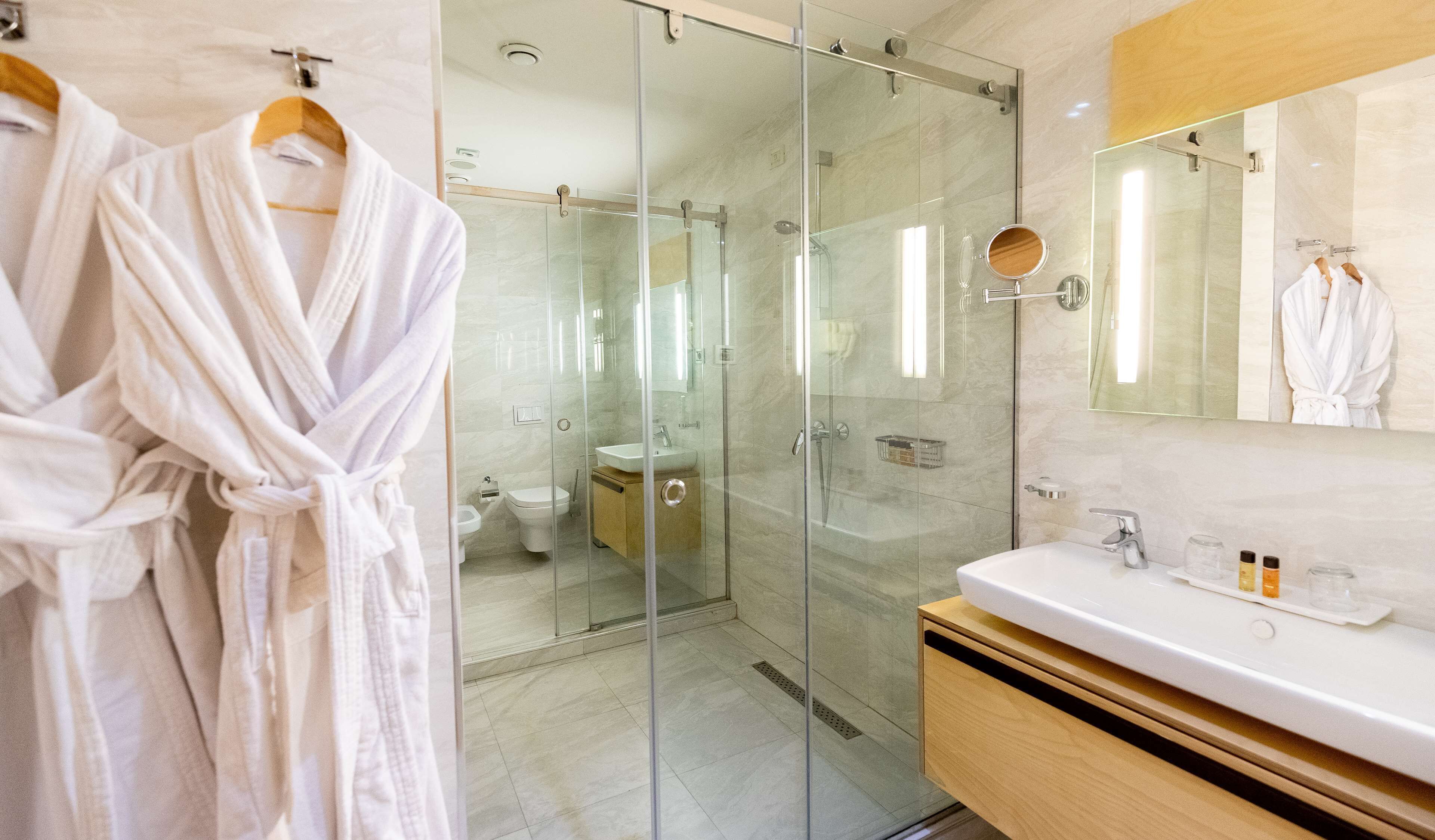 Un baño moderno con ducha de vidrio, tocador con espejo y dos batas blancas colgadas en un perchero.