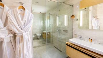 Un baño moderno con ducha de vidrio, tocador con espejo y dos batas blancas colgadas en un perchero.