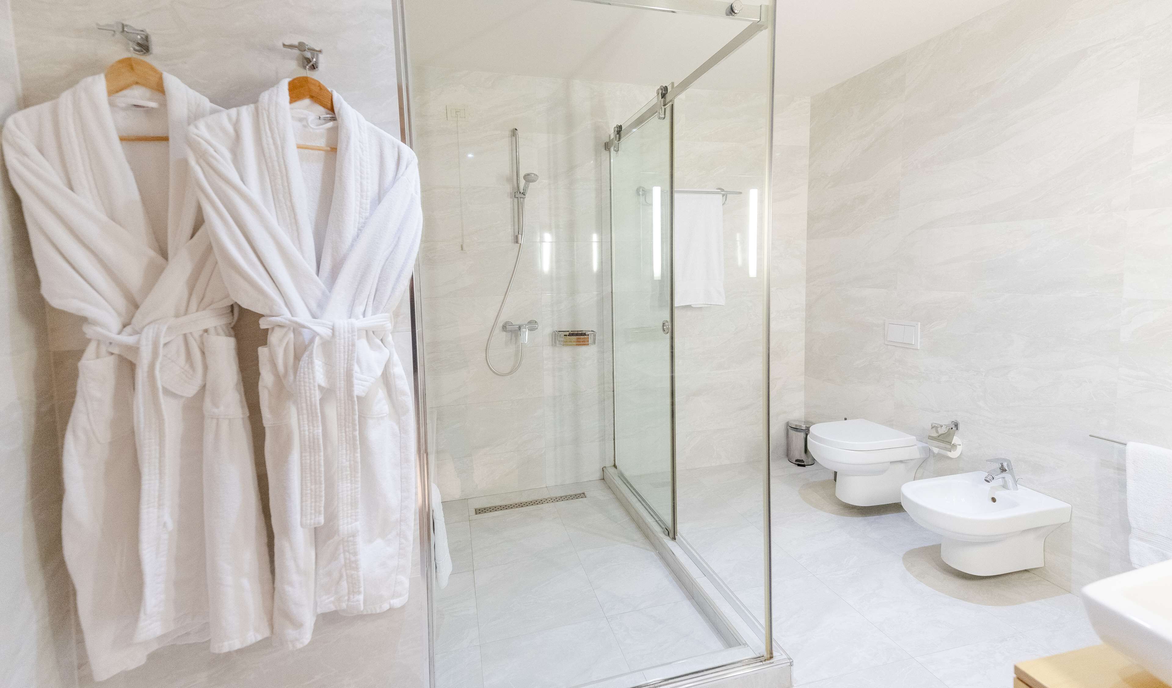 A modern bathroom with a glass-enclosed shower, a bidet, and two white robes hanging on a rack.