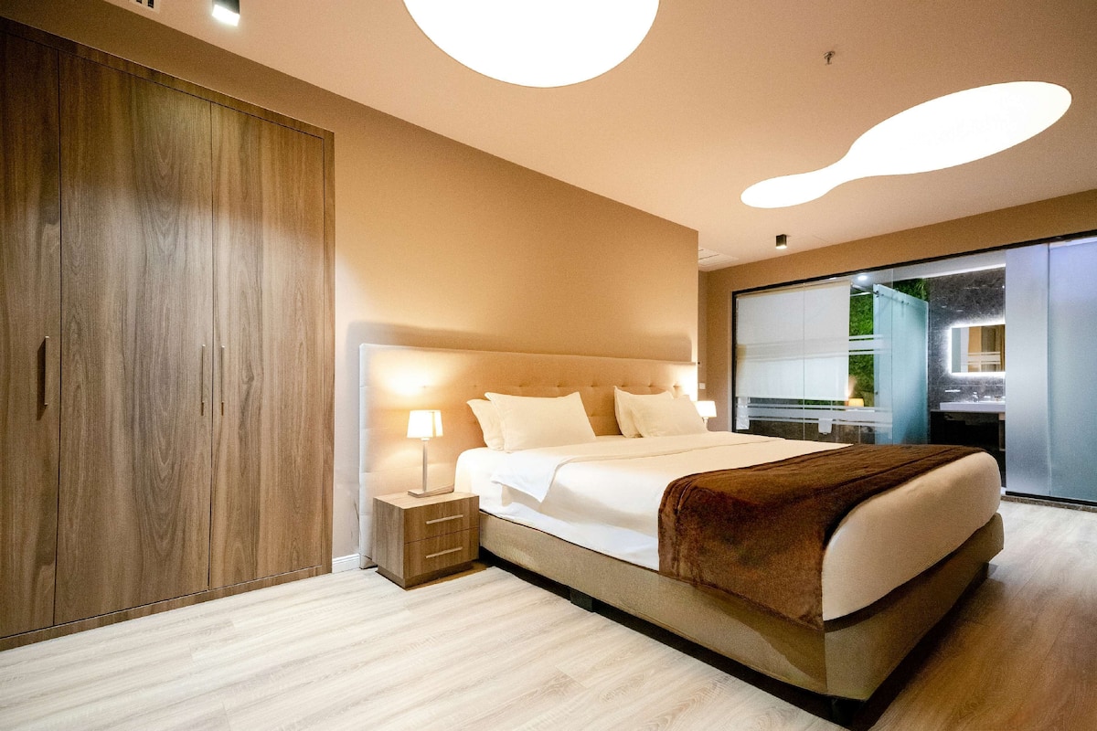 A modern bedroom with a large bed, wooden wardrobe, and a bathroom visible through a glass door.