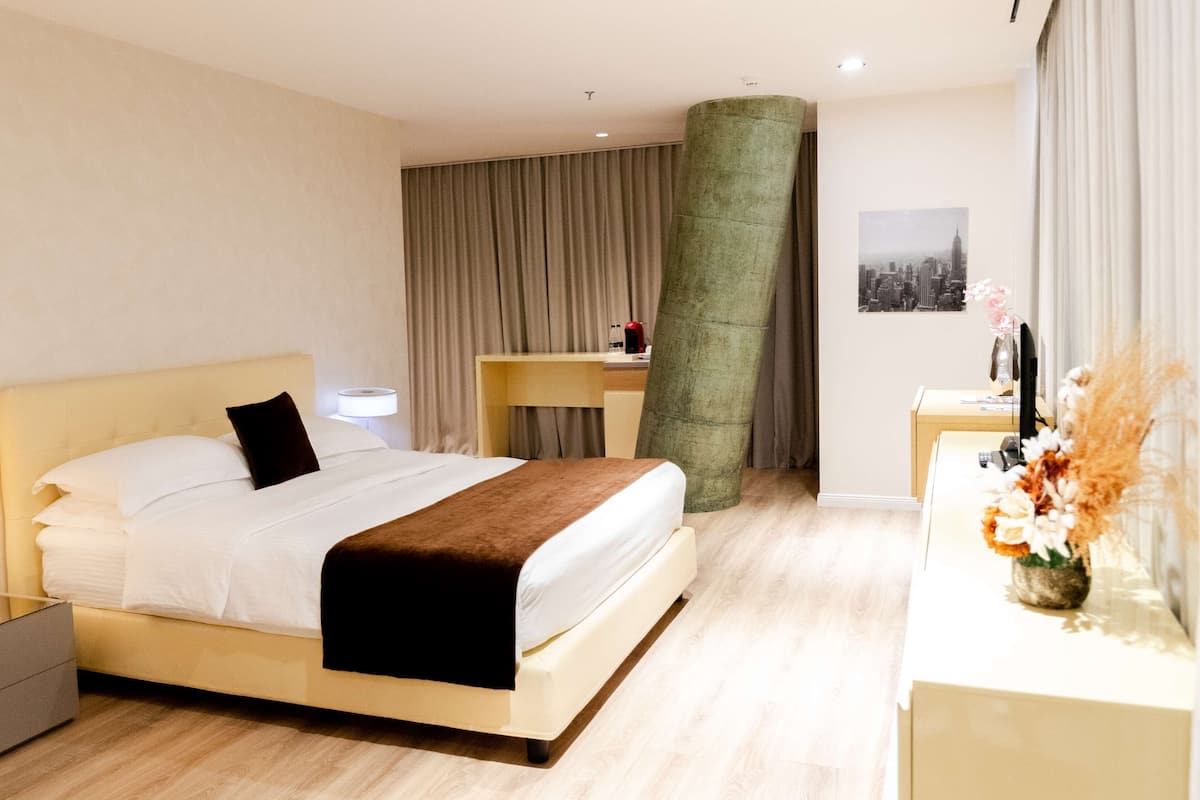 A hotel room with a bed, bedside table, lamp, desk, chair, and a large pillar.