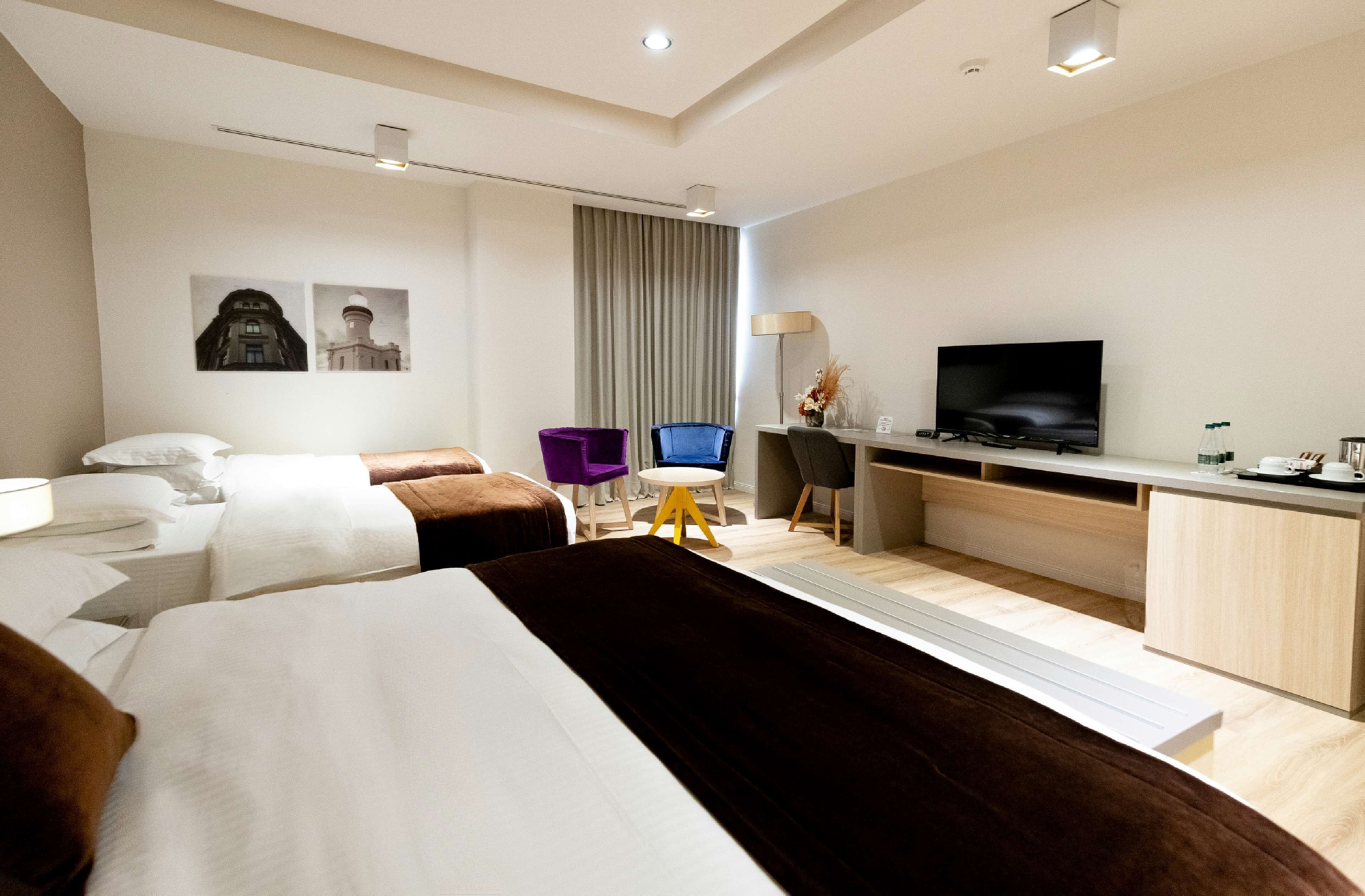 A hotel room with two beds, a desk with a TV, a chair, and a small table with a lamp.