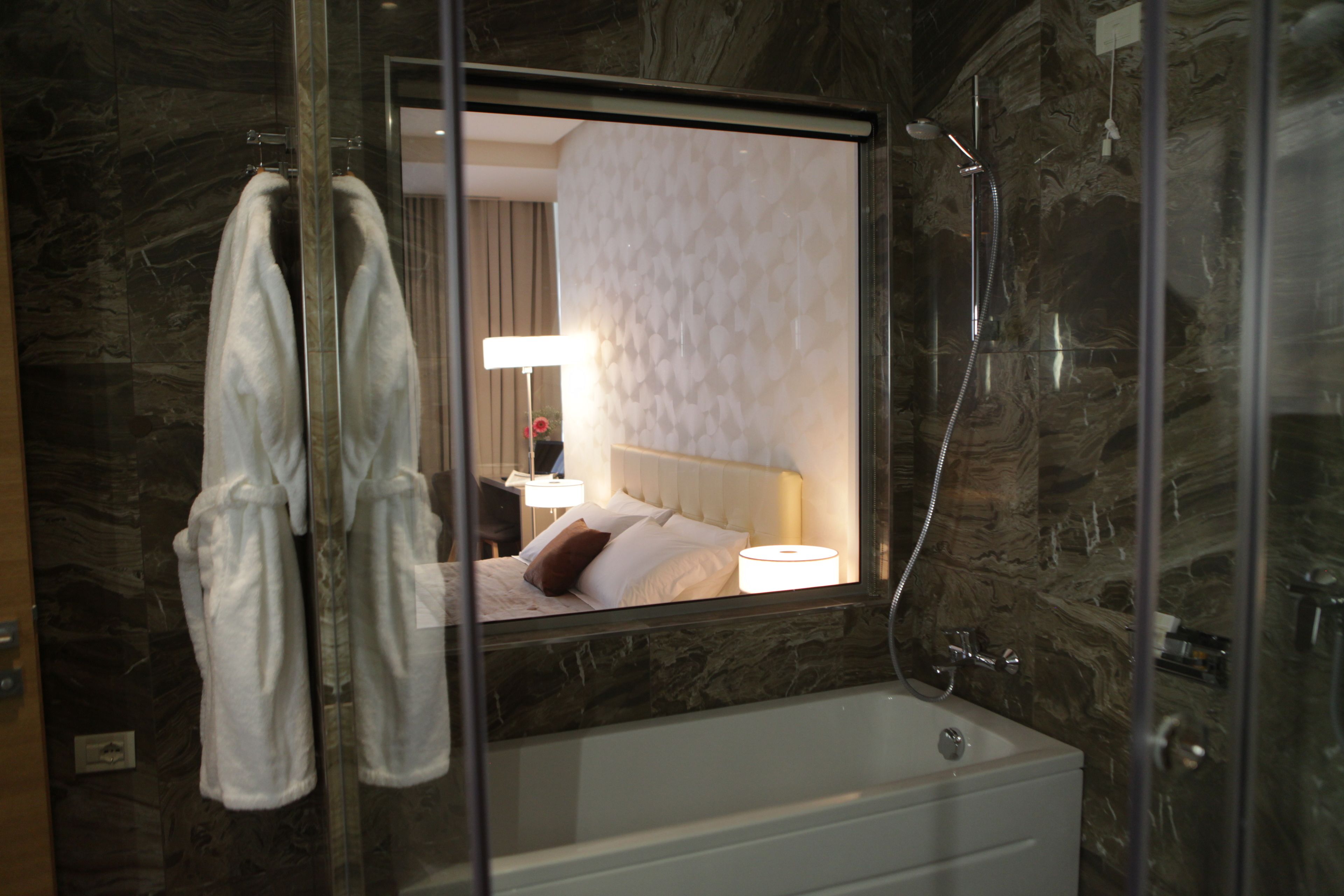 Un baño con cabina de ducha de vidrio, bañera y espejo que refleja una cama con una mesita de noche.