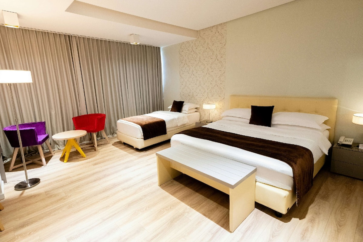 A hotel room with two beds, a desk, a chair, a bedside table, and a lamp.