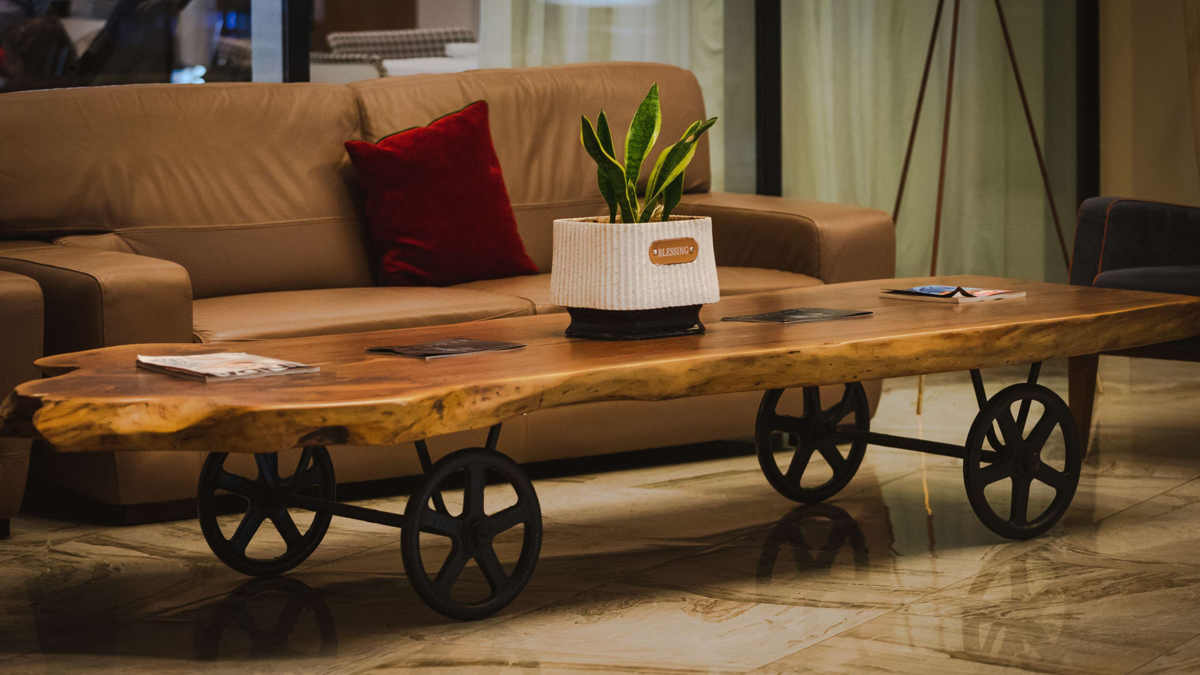 Una mesa de centro de madera con ruedas, una planta en maceta y un cojín rojo sobre un sofá de cuero en un interior moderno.