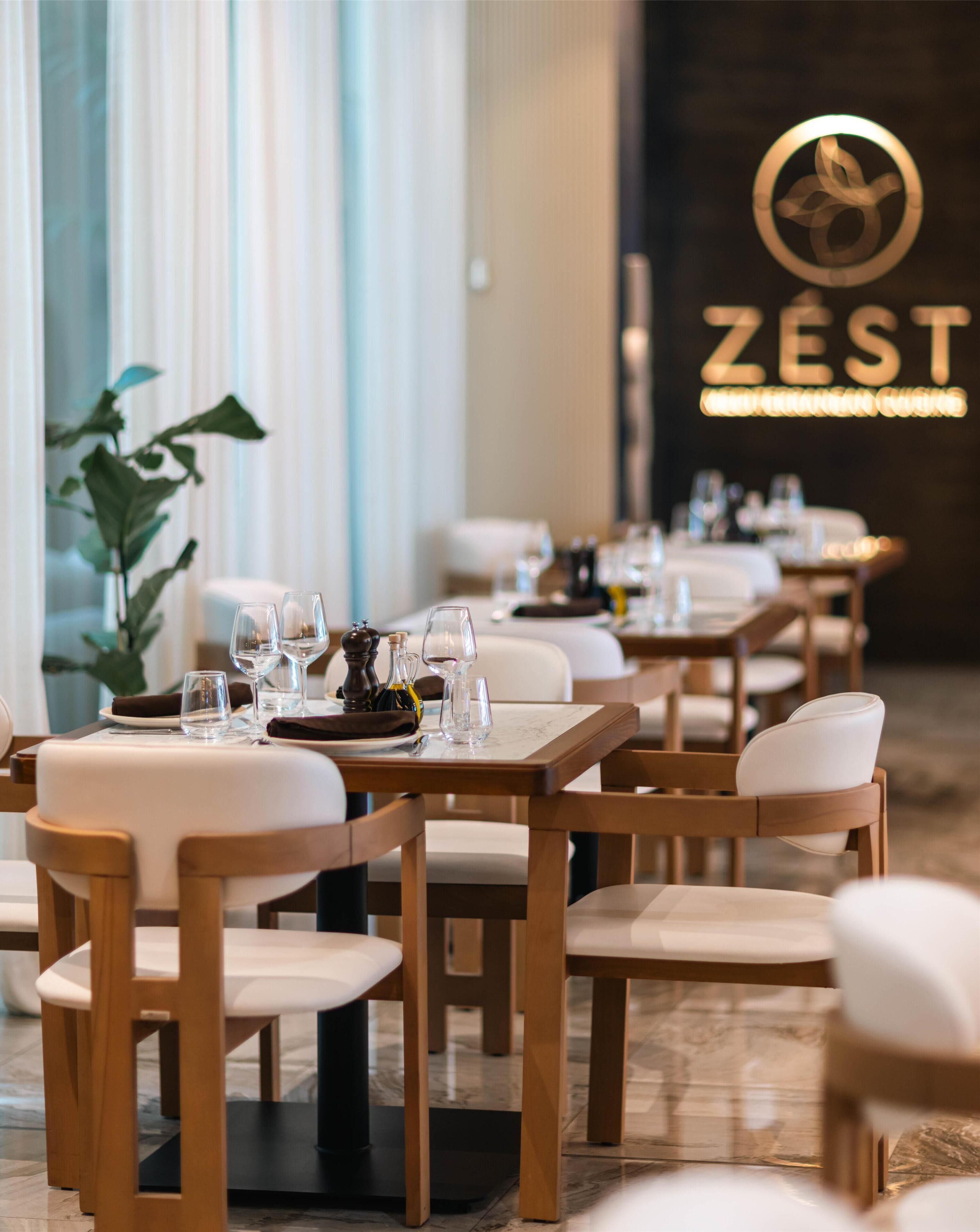 L’intérieur d’un restaurant avec des tables en bois et des chaises blanches, une plante et une enseigne sur laquelle on peut lire « ZEST ».