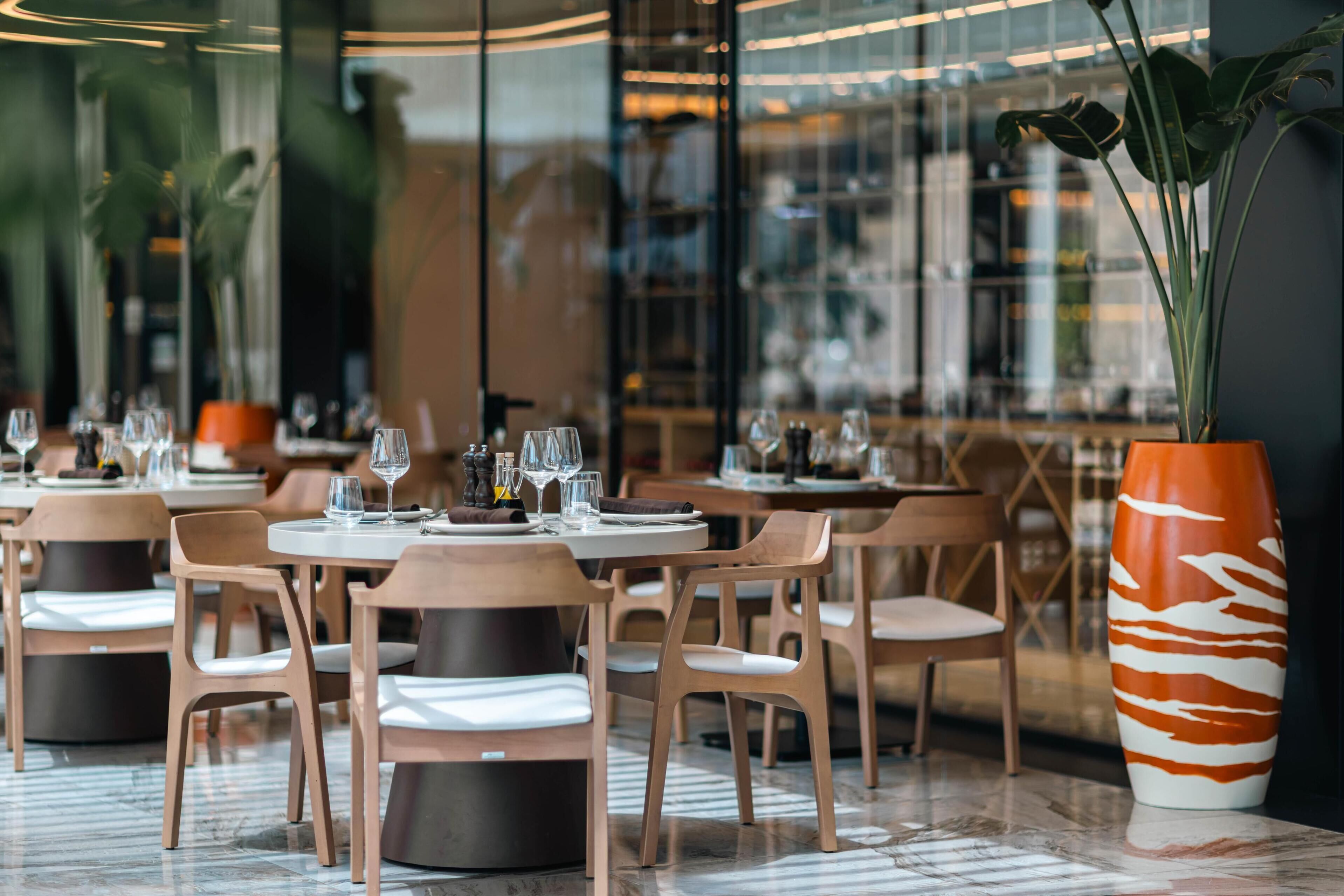 A restaurant with round tables, wooden chairs, and a large decorative vase.