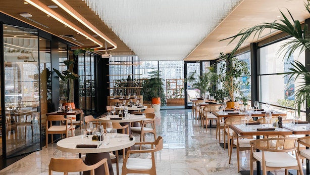 Un restaurant moderne, avec un intérieur agrémenté de tables rondes, de chaises en bois et de grandes fenêtres offrant une vue sur la ville.