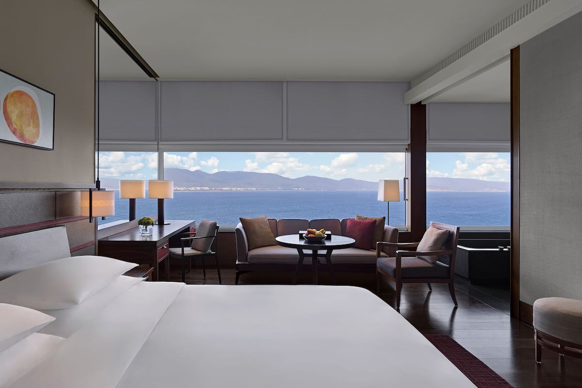 A hotel room with a large bed, a seating area with a table, and a view of the sea and mountains.