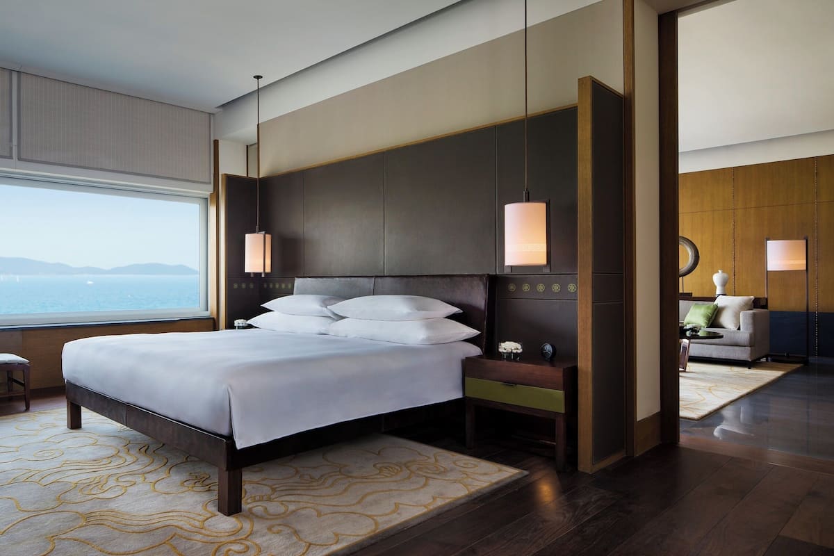 A modern hotel room with a large bed, a view of the sea, a sitting area with a chair, and a nightstand with a lamp.