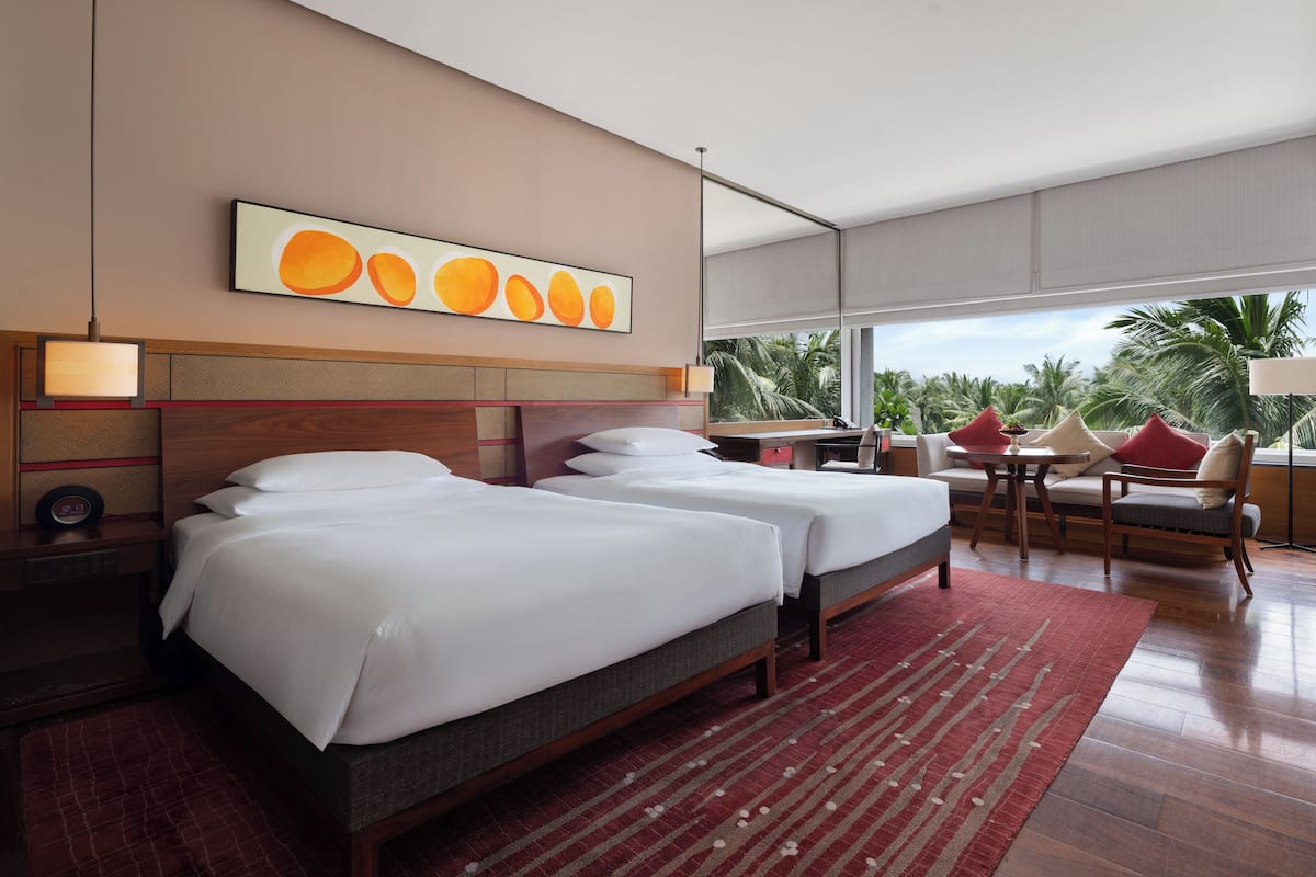 A hotel room with two beds, a red rug, a large window with a view of palm trees, and a wall art piece.